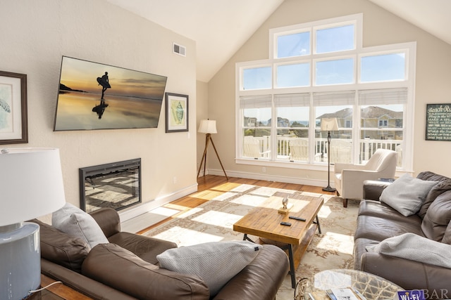 living area featuring light wood finished floors, visible vents, a fireplace with flush hearth, high vaulted ceiling, and baseboards