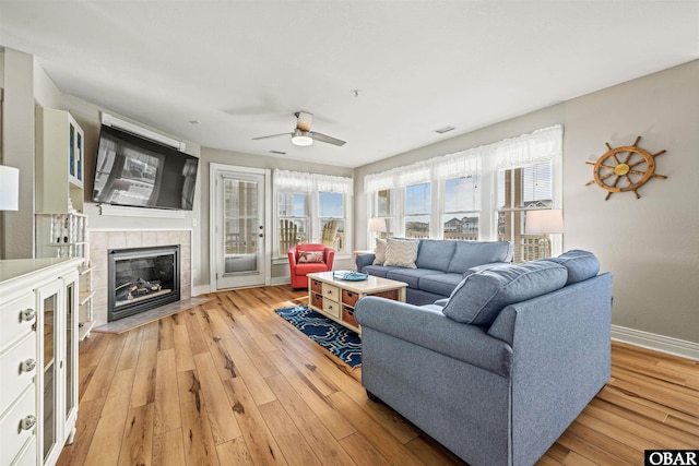 living area featuring visible vents, a tiled fireplace, a ceiling fan, light wood-type flooring, and baseboards