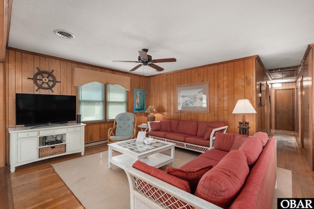 living area featuring visible vents, baseboard heating, a ceiling fan, wooden walls, and wood finished floors