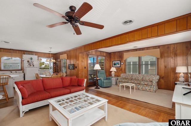 living area featuring visible vents, wood walls, light wood-style flooring, and ceiling fan with notable chandelier