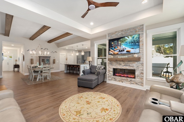 living room featuring baseboards, beamed ceiling, wood finished floors, a stone fireplace, and ceiling fan with notable chandelier