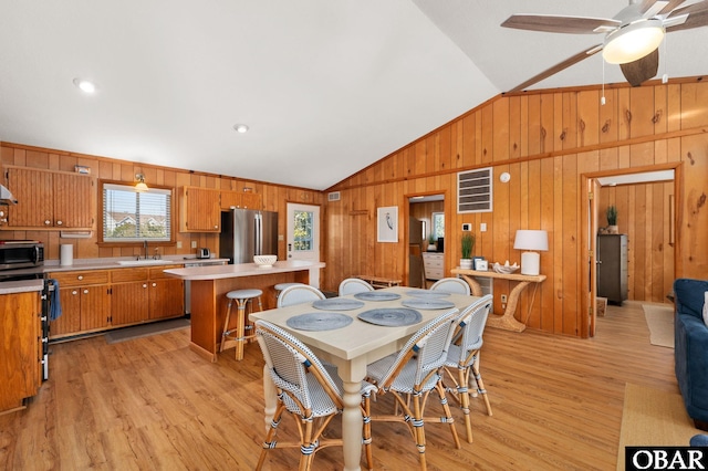 dining room featuring a ceiling fan, vaulted ceiling, wooden walls, and light wood-style floors