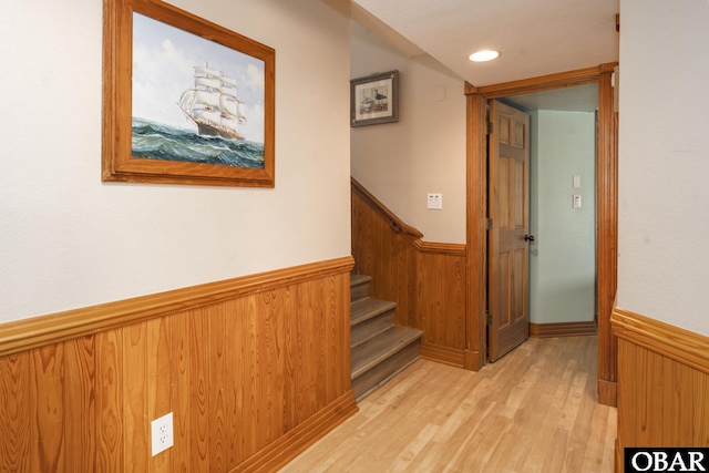 hallway with wooden walls, recessed lighting, light wood-style floors, stairs, and wainscoting