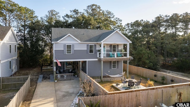 back of property featuring a shingled roof, concrete driveway, a balcony, a fenced backyard, and a carport