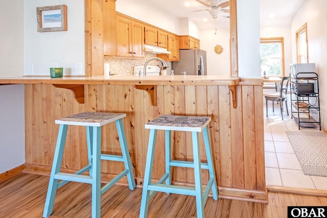 kitchen with tasteful backsplash, a ceiling fan, a kitchen breakfast bar, under cabinet range hood, and stainless steel refrigerator with ice dispenser