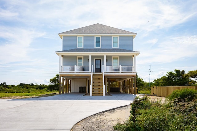 raised beach house with a shingled roof, covered porch, concrete driveway, stairway, and a carport