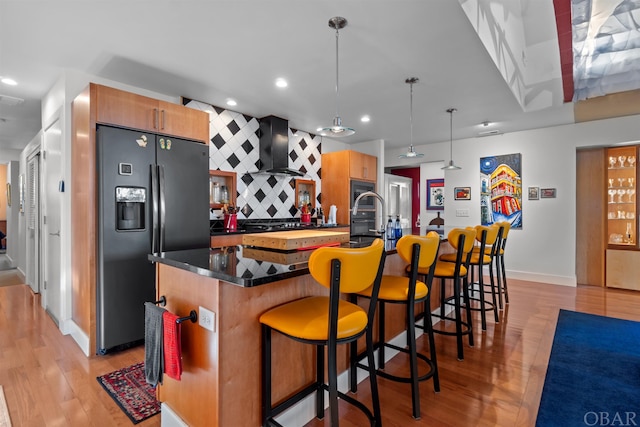 kitchen with decorative light fixtures, dark countertops, black fridge with ice dispenser, a kitchen island with sink, and wall chimney range hood