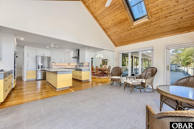 kitchen with a skylight, light countertops, a kitchen island with sink, stainless steel fridge, and wall chimney exhaust hood