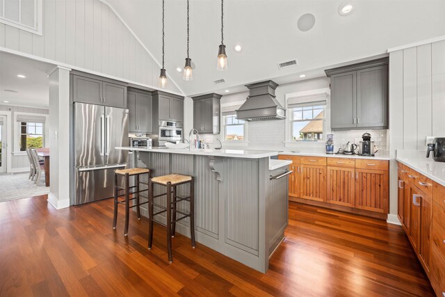 kitchen featuring brown cabinets, custom exhaust hood, stainless steel appliances, light countertops, and a kitchen island with sink