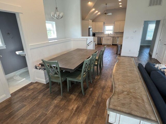 dining space with high vaulted ceiling, ceiling fan with notable chandelier, dark wood-style flooring, visible vents, and wainscoting