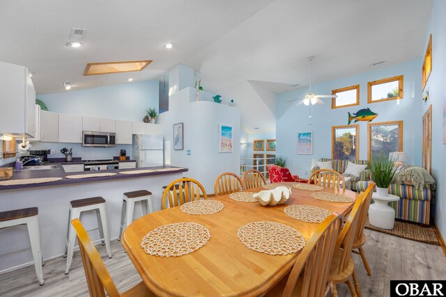 dining space with ceiling fan, high vaulted ceiling, a skylight, and light wood-style floors