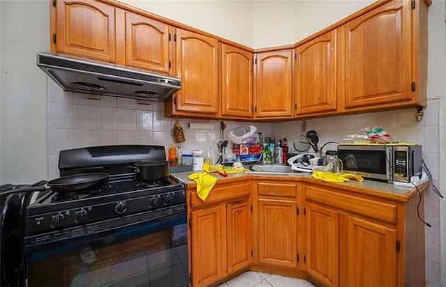 kitchen with stainless steel microwave, black gas range, backsplash, brown cabinetry, and under cabinet range hood