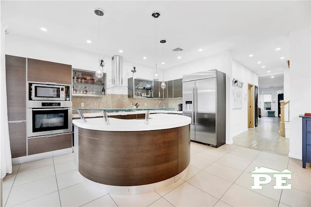 kitchen with pendant lighting, wall chimney exhaust hood, decorative backsplash, an island with sink, and appliances with stainless steel finishes