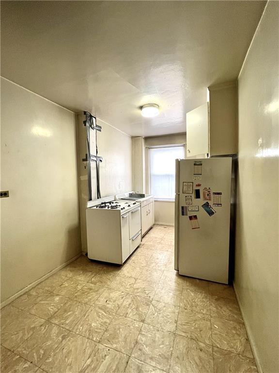 kitchen featuring light floors, baseboards, white range with gas stovetop, freestanding refrigerator, and white cabinets