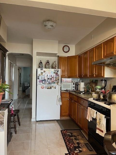 kitchen with brown cabinets, under cabinet range hood, white appliances, light tile patterned floors, and decorative backsplash