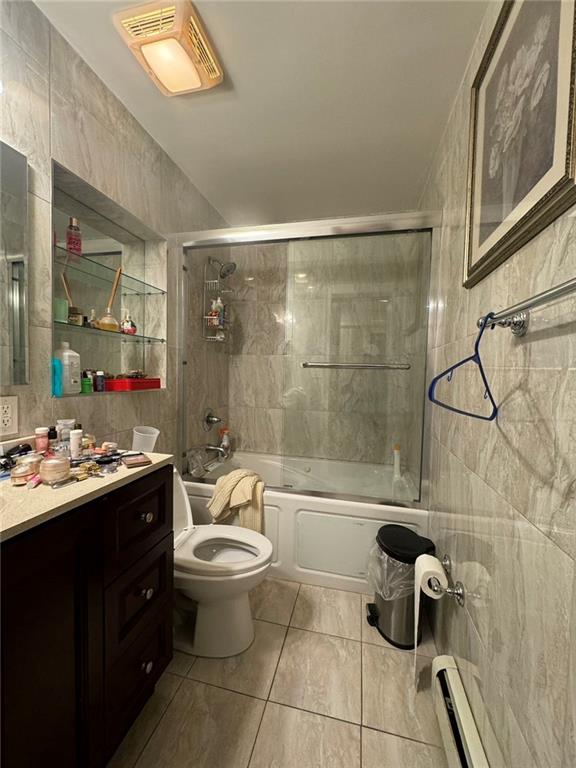 full bathroom with toilet, shower / bath combination with glass door, tile walls, vanity, and a baseboard heating unit