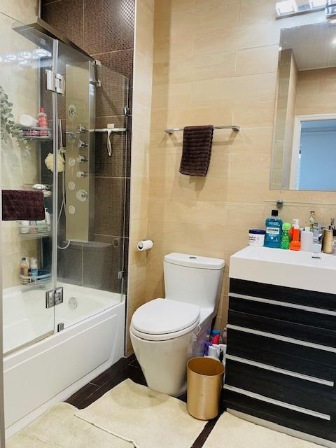 full bathroom featuring bath / shower combo with glass door, tile walls, vanity, and toilet