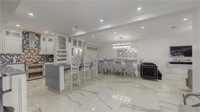 kitchen with tasteful backsplash, white cabinets, a breakfast bar area, double oven range, and wall chimney range hood