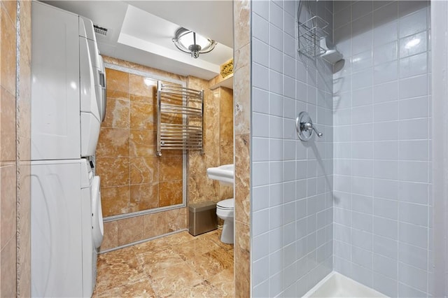 bathroom with a tile shower, stacked washer and dryer, radiator heating unit, and toilet