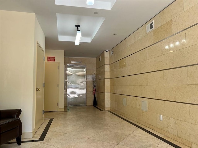 hall with light tile patterned floors, tile walls, and a tray ceiling