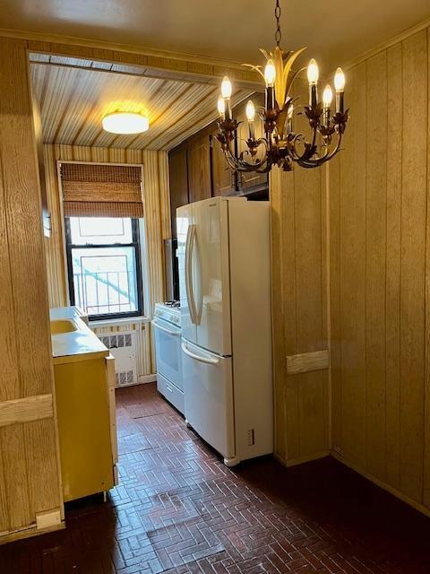 kitchen featuring white appliances, wooden walls, radiator, brick floor, and a chandelier