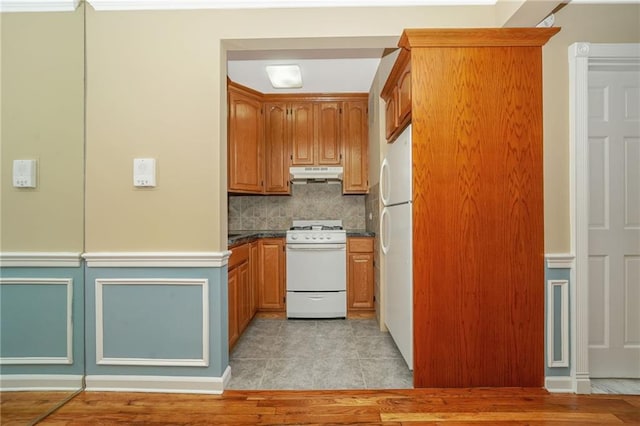 kitchen featuring dark countertops, under cabinet range hood, decorative backsplash, brown cabinetry, and white appliances