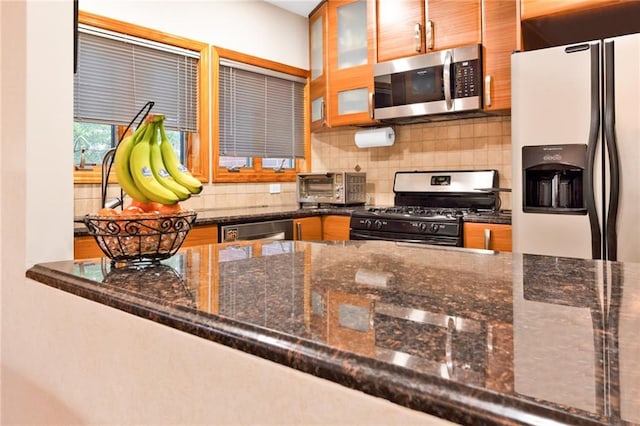 kitchen with a toaster, stainless steel appliances, decorative backsplash, dark stone countertops, and glass insert cabinets