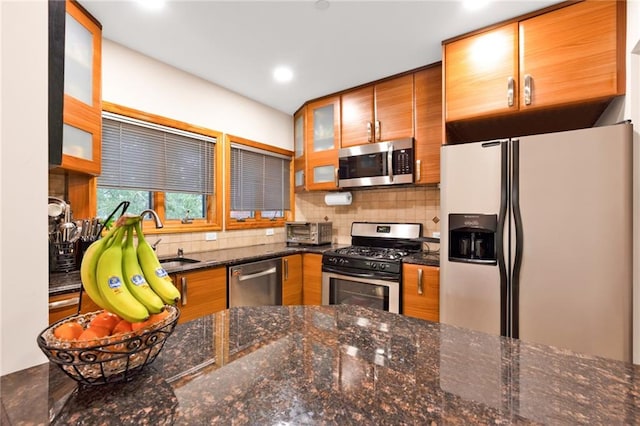 kitchen with decorative backsplash, glass insert cabinets, brown cabinets, dark stone countertops, and stainless steel appliances