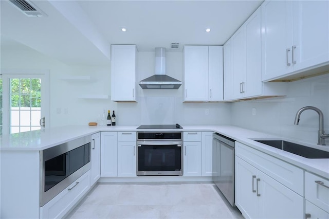 kitchen featuring kitchen peninsula, appliances with stainless steel finishes, white cabinetry, wall chimney exhaust hood, and sink