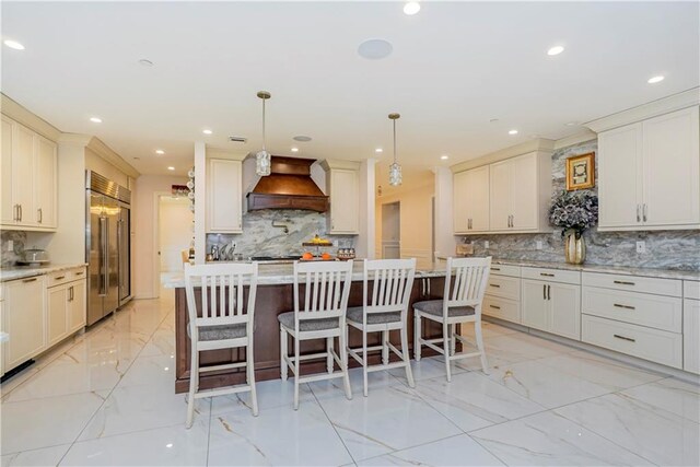 kitchen with hanging light fixtures, a kitchen breakfast bar, stainless steel built in refrigerator, an island with sink, and custom exhaust hood