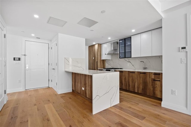 kitchen featuring white cabinetry, backsplash, light hardwood / wood-style floors, light stone countertops, and wall chimney exhaust hood