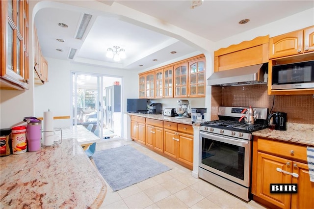 kitchen featuring wall chimney range hood, light tile patterned floors, appliances with stainless steel finishes, a tray ceiling, and light stone countertops