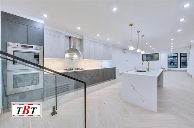 kitchen with light parquet floors, sink, wall chimney range hood, decorative light fixtures, and a center island with sink