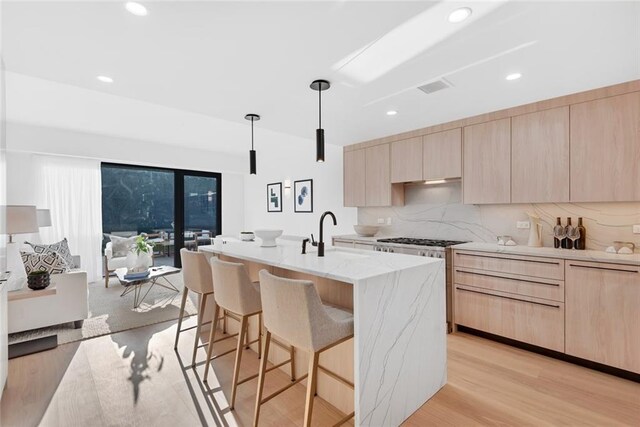 kitchen with an island with sink, hanging light fixtures, light hardwood / wood-style flooring, light brown cabinetry, and light stone counters