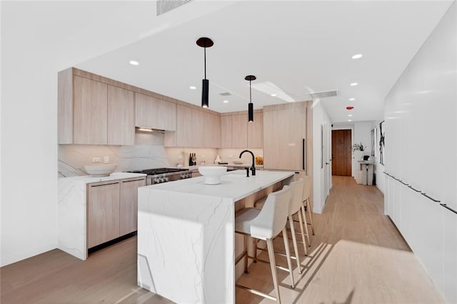 kitchen featuring a center island with sink, light stone countertops, light brown cabinetry, light hardwood / wood-style floors, and decorative light fixtures