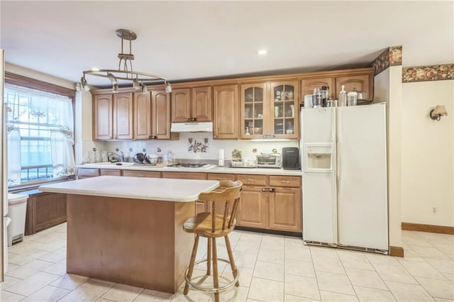 kitchen with white refrigerator with ice dispenser, stainless steel gas stovetop, a kitchen island, sink, and decorative light fixtures