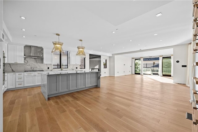 kitchen with wall chimney exhaust hood, open floor plan, light wood-type flooring, white cabinetry, and backsplash