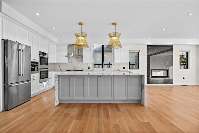 kitchen featuring a fireplace, stainless steel appliances, light countertops, white cabinetry, and wall chimney range hood