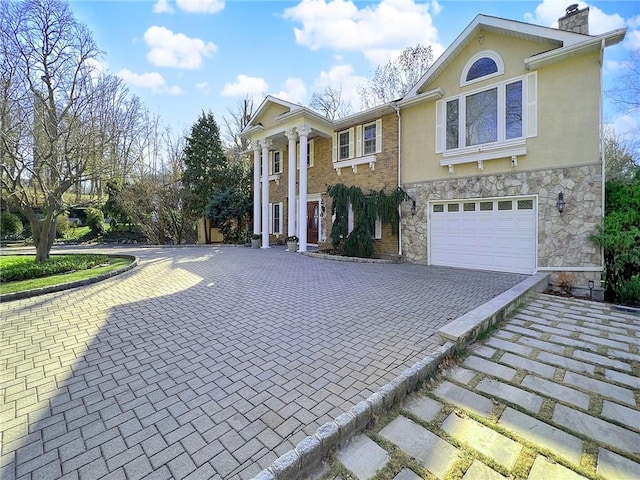 greek revival inspired property featuring stucco siding, a chimney, stone siding, decorative driveway, and a garage