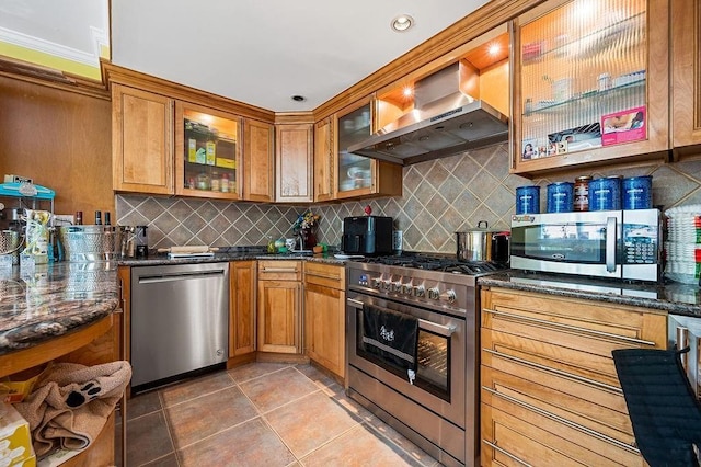 kitchen featuring appliances with stainless steel finishes, backsplash, wall chimney exhaust hood, dark stone countertops, and glass insert cabinets