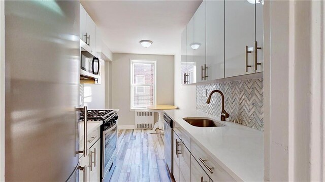 kitchen with stainless steel appliances, sink, radiator heating unit, light hardwood / wood-style floors, and white cabinetry