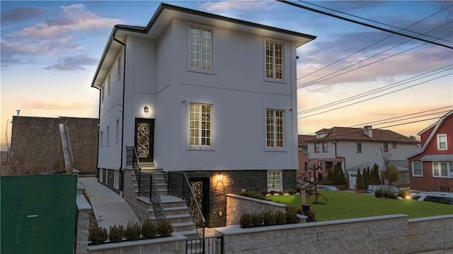 front of property at dusk featuring a yard, stone siding, and stucco siding