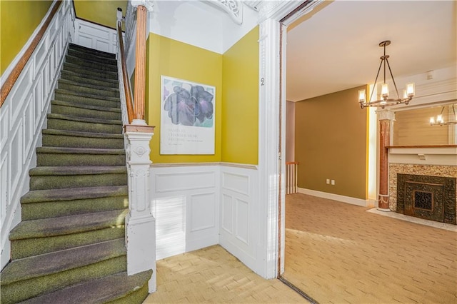 stairs with an inviting chandelier, a fireplace with flush hearth, a decorative wall, and wainscoting