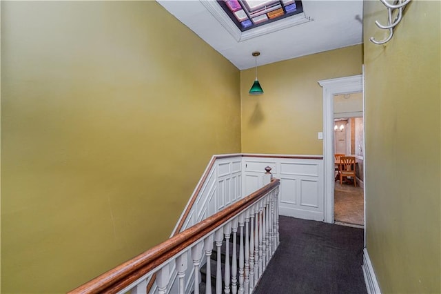 hallway featuring a decorative wall, a wainscoted wall, a skylight, an upstairs landing, and dark carpet