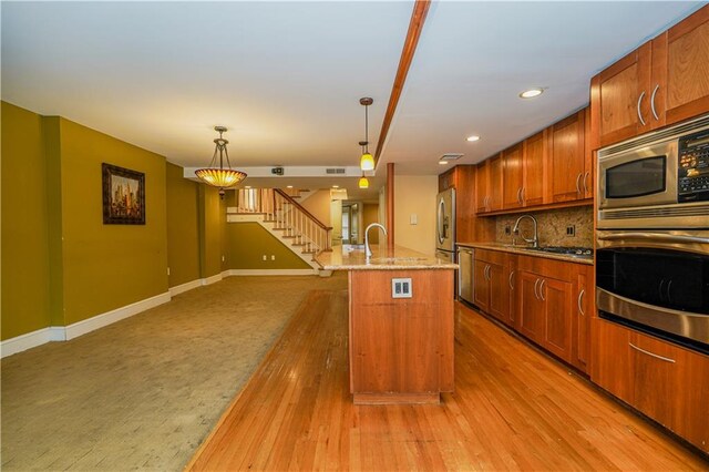 kitchen with appliances with stainless steel finishes, an island with sink, light stone countertops, light hardwood / wood-style flooring, and decorative light fixtures