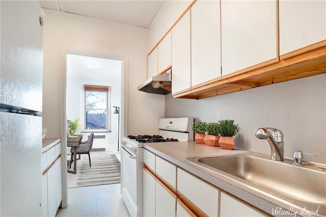 kitchen with white gas stove, under cabinet range hood, freestanding refrigerator, white cabinetry, and a sink