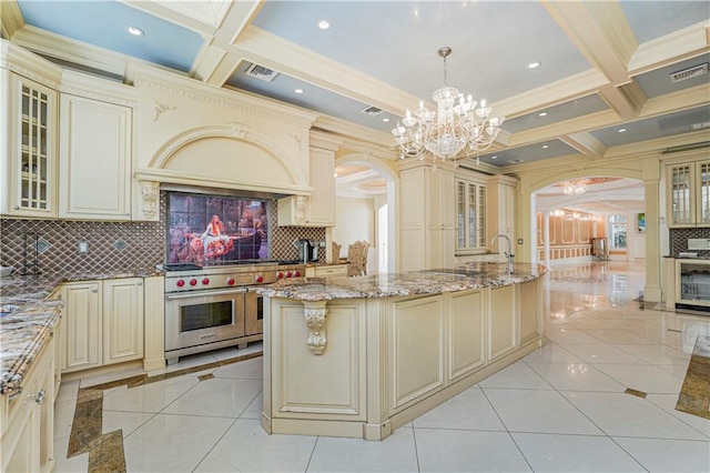 kitchen featuring cream cabinets, light tile patterned floors, pendant lighting, range with two ovens, and an island with sink