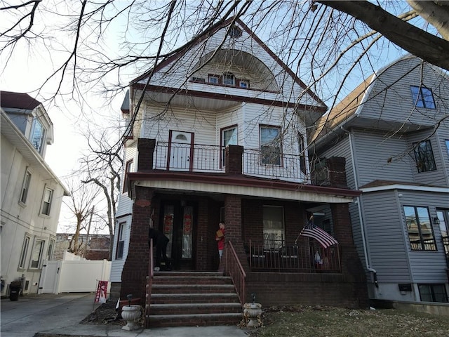 victorian house featuring brick siding, a porch, a balcony, and fence