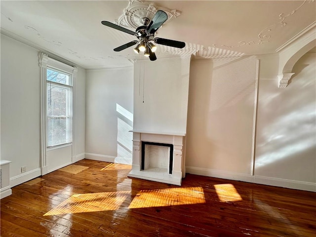 unfurnished living room featuring a fireplace with raised hearth, hardwood / wood-style floors, ornamental molding, a ceiling fan, and baseboards