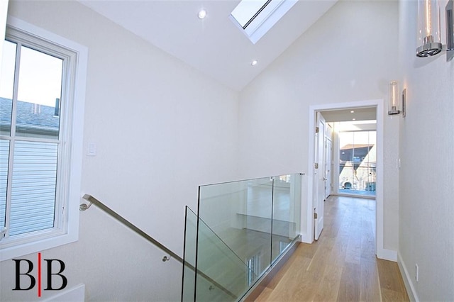 corridor featuring a skylight, high vaulted ceiling, baseboards, an upstairs landing, and light wood-style flooring